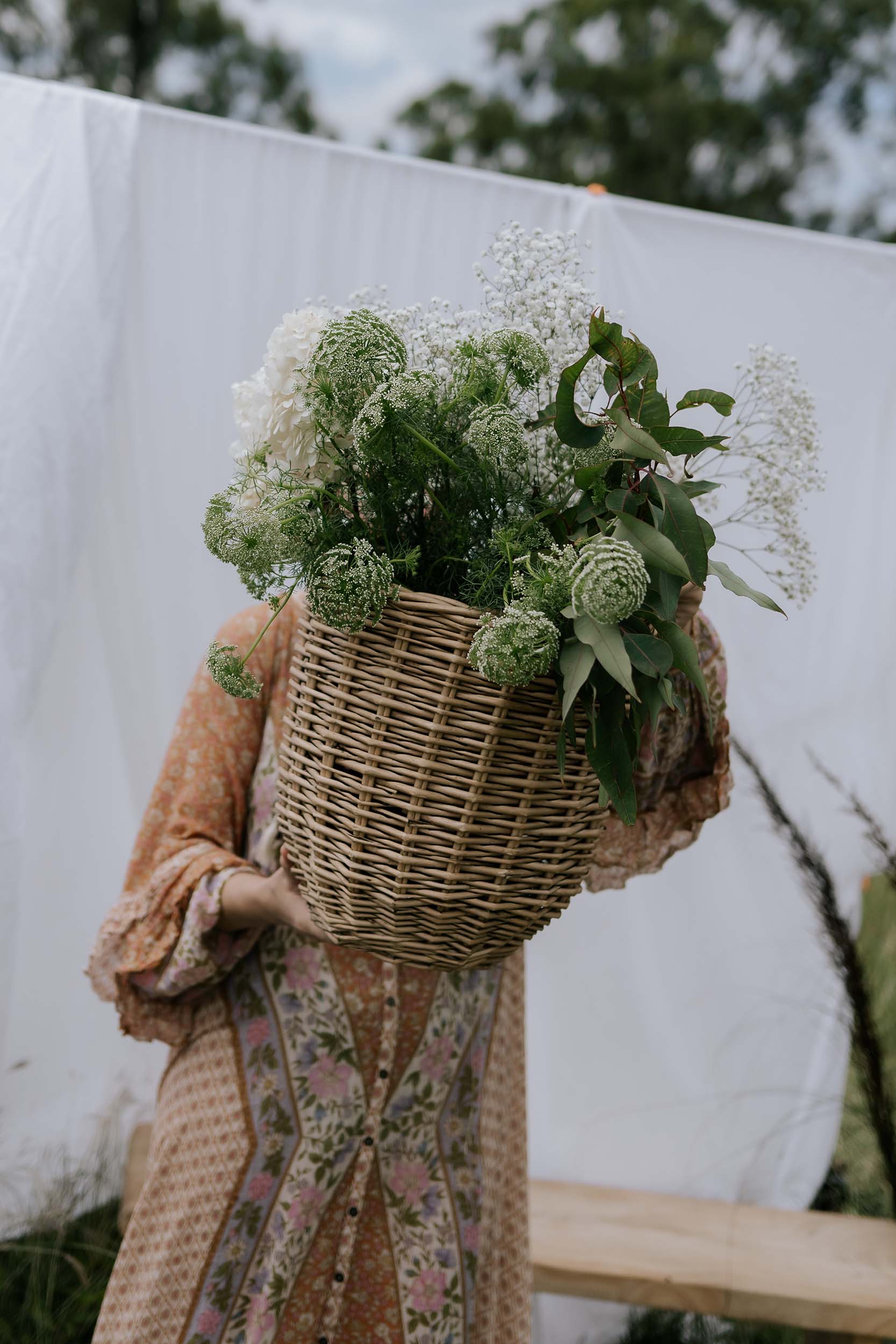 Person holding a large woven basket filled with greenery against a white curtain background